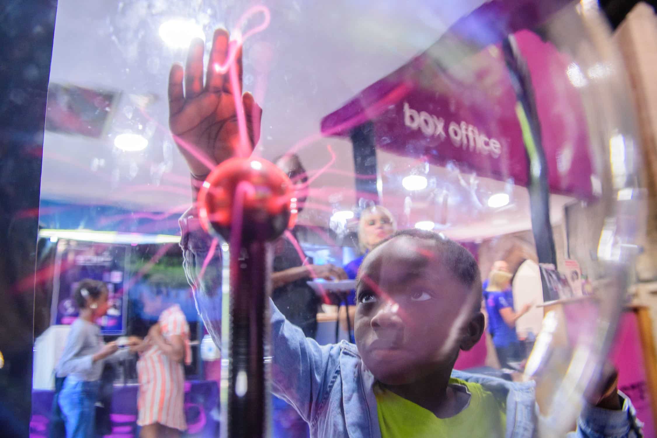 A child interacts with a plasma ball, with people and a "box office" sign visible in the background.
