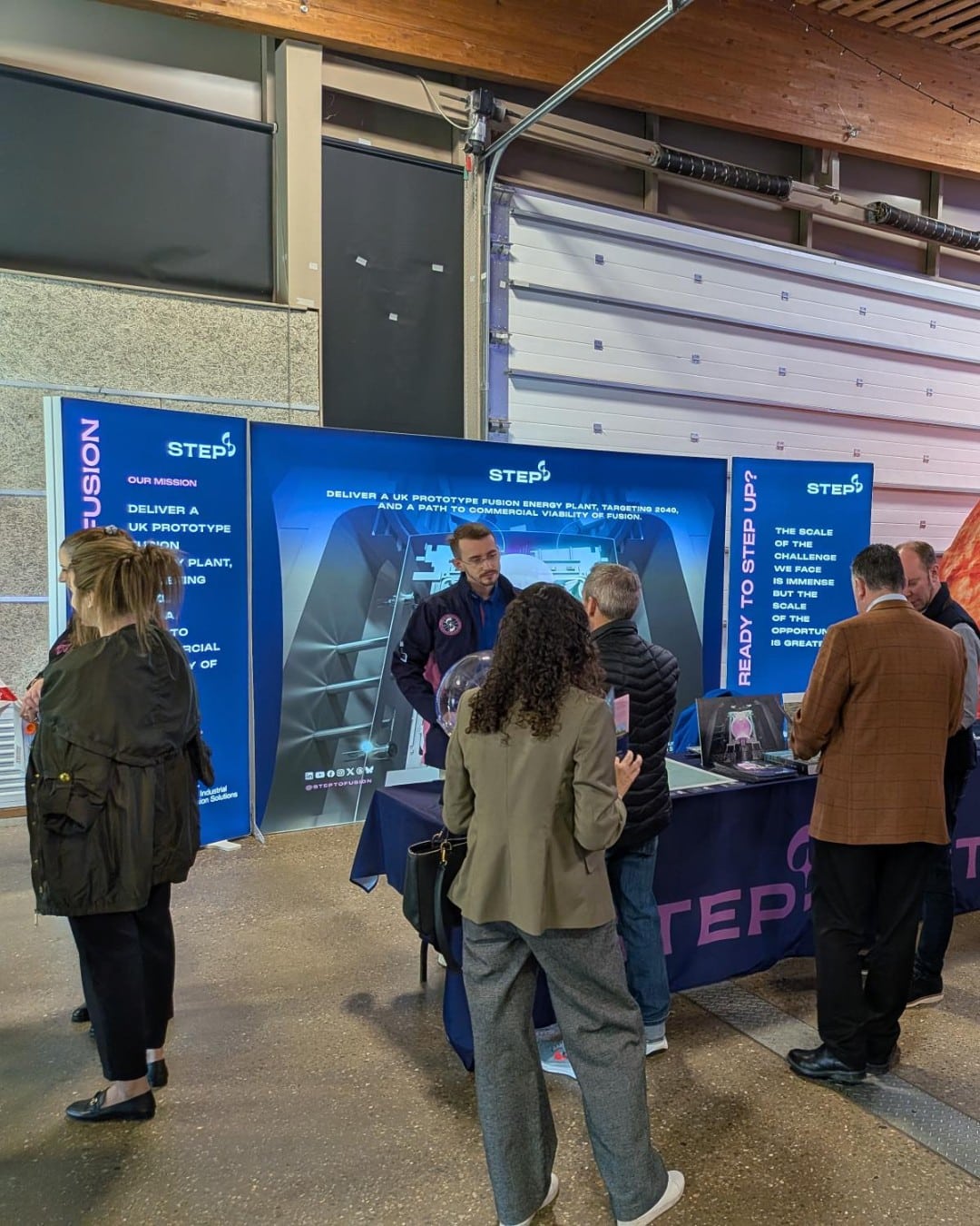 STEP Fusion branded exhibitions stand, explaining what the STEP programme is. Male member of STEP team is talking to members of the public about fusion.